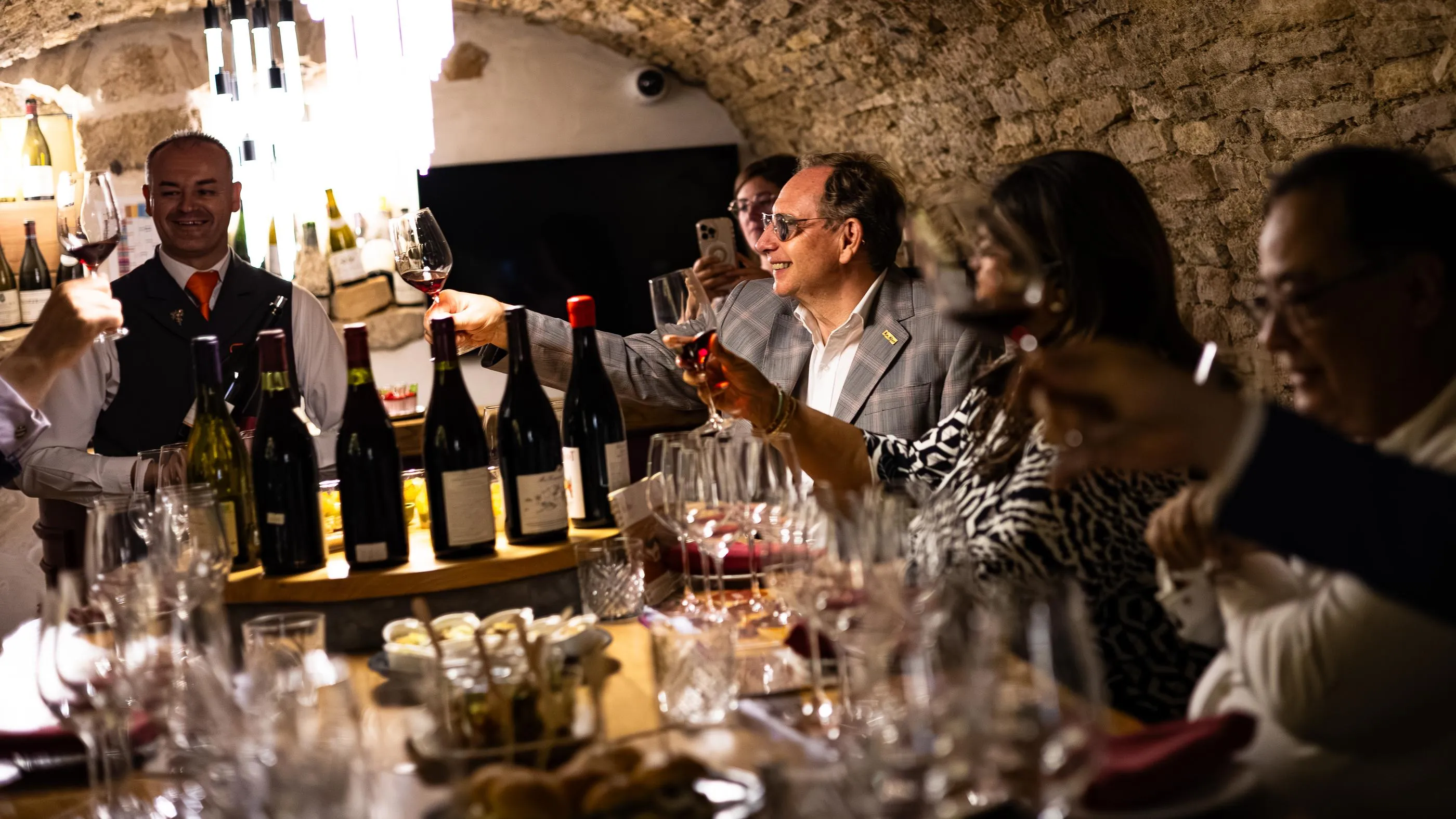 Un groupe de personnes participe à une dégustation de vin dans une cave, entourées de bouteilles et de verres à vin