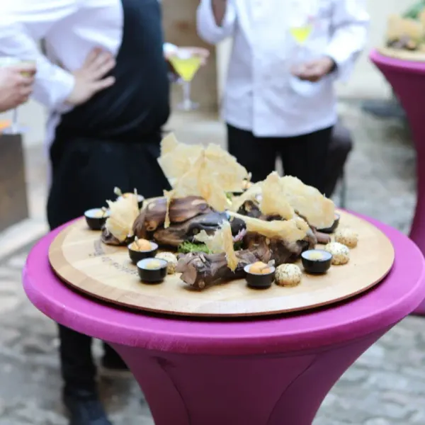 Un plat de viande accompagné de garnitures et de sauces est posé sur une table haute violette, entouré de personnes en tenue formelle tenant des verres