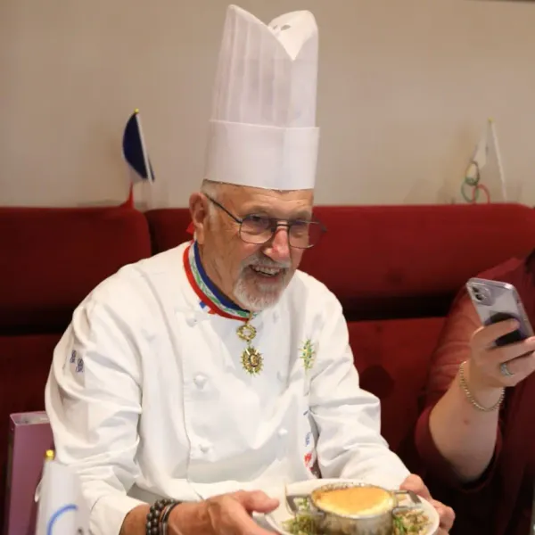 Un chef en uniforme blanc avec un haut chapeau et des médailles présente un plat à une personne tenant un smartphone
