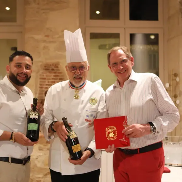 Trois hommes posent ensemble dans une pièce, l'un en tenue de chef avec une médaille, tenant des bouteilles de vin, l'autre un livre rouge