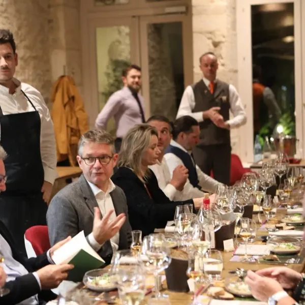 Un groupe de personnes assises autour d'une table avec des verres et des plats, un chef en tablier noir debout à gauche