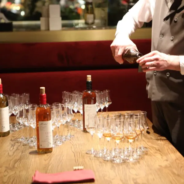 Un serveur en veste verse du vin dans des verres alignés sur un comptoir en bois, avec plusieurs bouteilles de vin à proximité