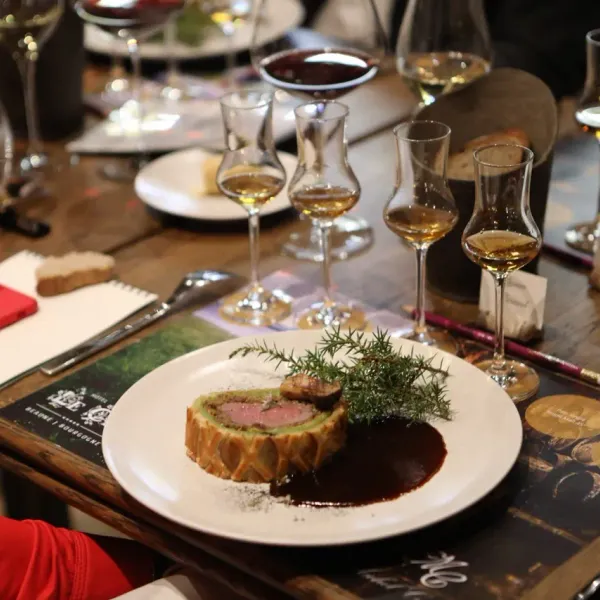 Un plat de viande en croûte avec une sauce et des herbes, entouré de verres de vin et d'ustensiles sur une table en bois