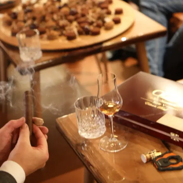 Une personne tient un cigare allumé près d'une table avec des verres de vin, des chocolats et une bouteille