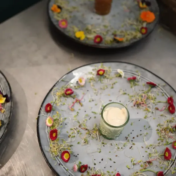 Trois assiettes transparentes avec des micro-pousses, des fleurs comestibles et des petits pots de crème au centre, posées sur une surface métallique