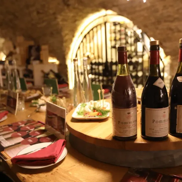 Bouteilles de vin Pomard sur une table en bois dans une cave à vin éclairée, accompagnées de plats et de couverts