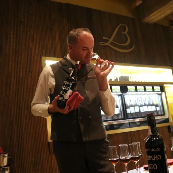 Un homme en costume verse du vin dans un verre dans un cadre intérieur avec des bouteilles et des verres sur une table en bois