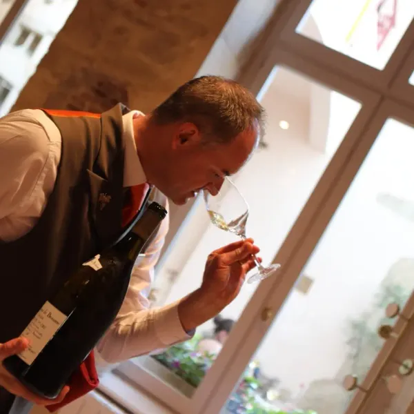 Un homme en veste et gilet examine un verre de vin, tenant une bouteille à la main, près d'une fenêtre ouverte